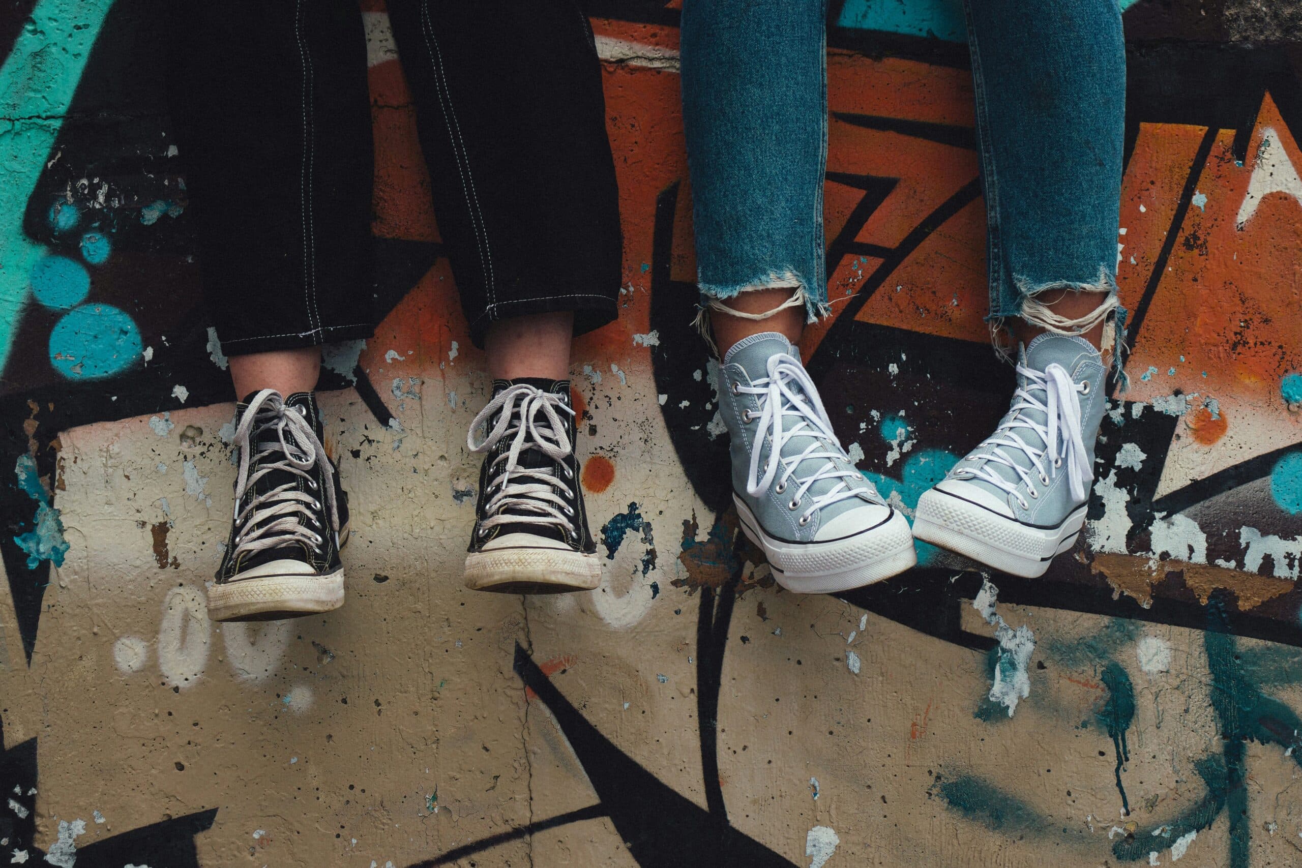 Two teenagers sitting on a graffitied wall. Showcasing their black and baby blue converse sneakers.